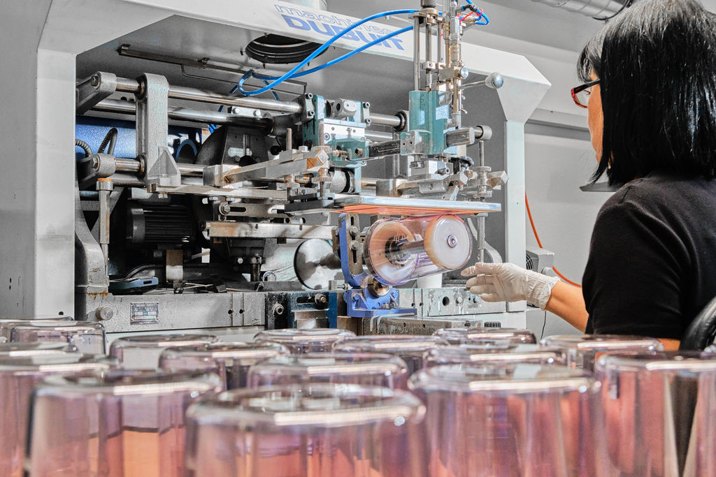 A woman wearing gloves operates a machine in a factory, with rows of clear plastic cups in the foreground and mechanical parts visible in the background.