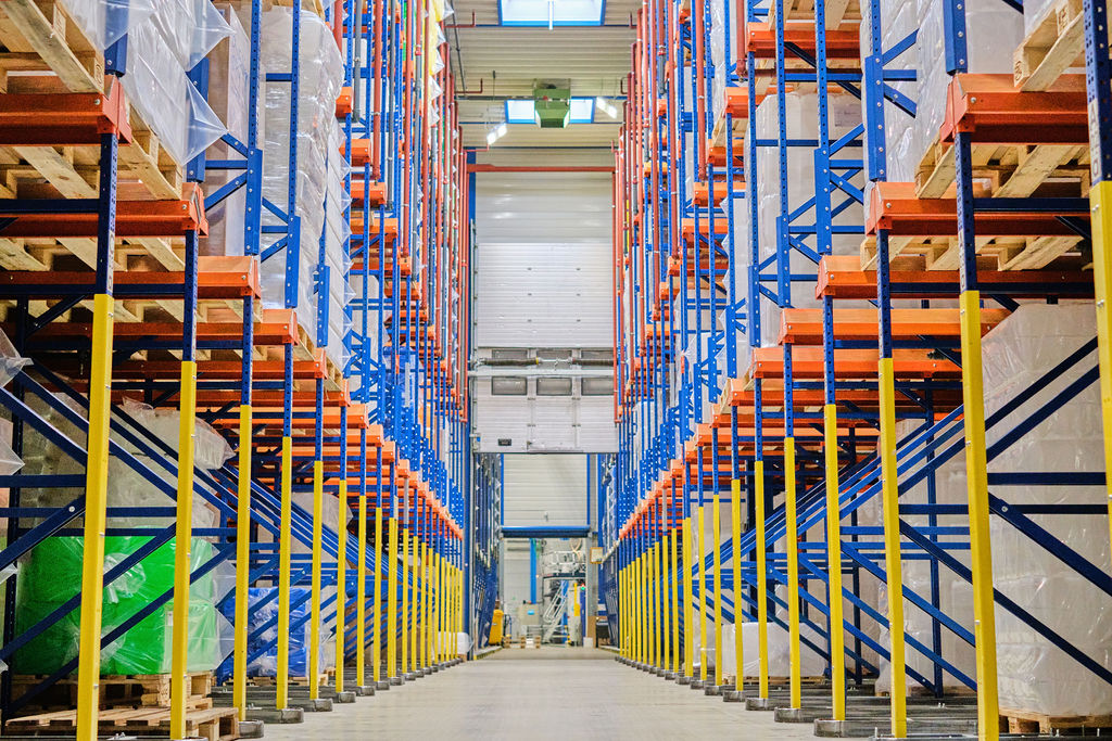 Warehouse interior with tall racks of pallets stacked on both sides of a wide central aisle, bright lighting, and yellow safety barriers along the shelves.